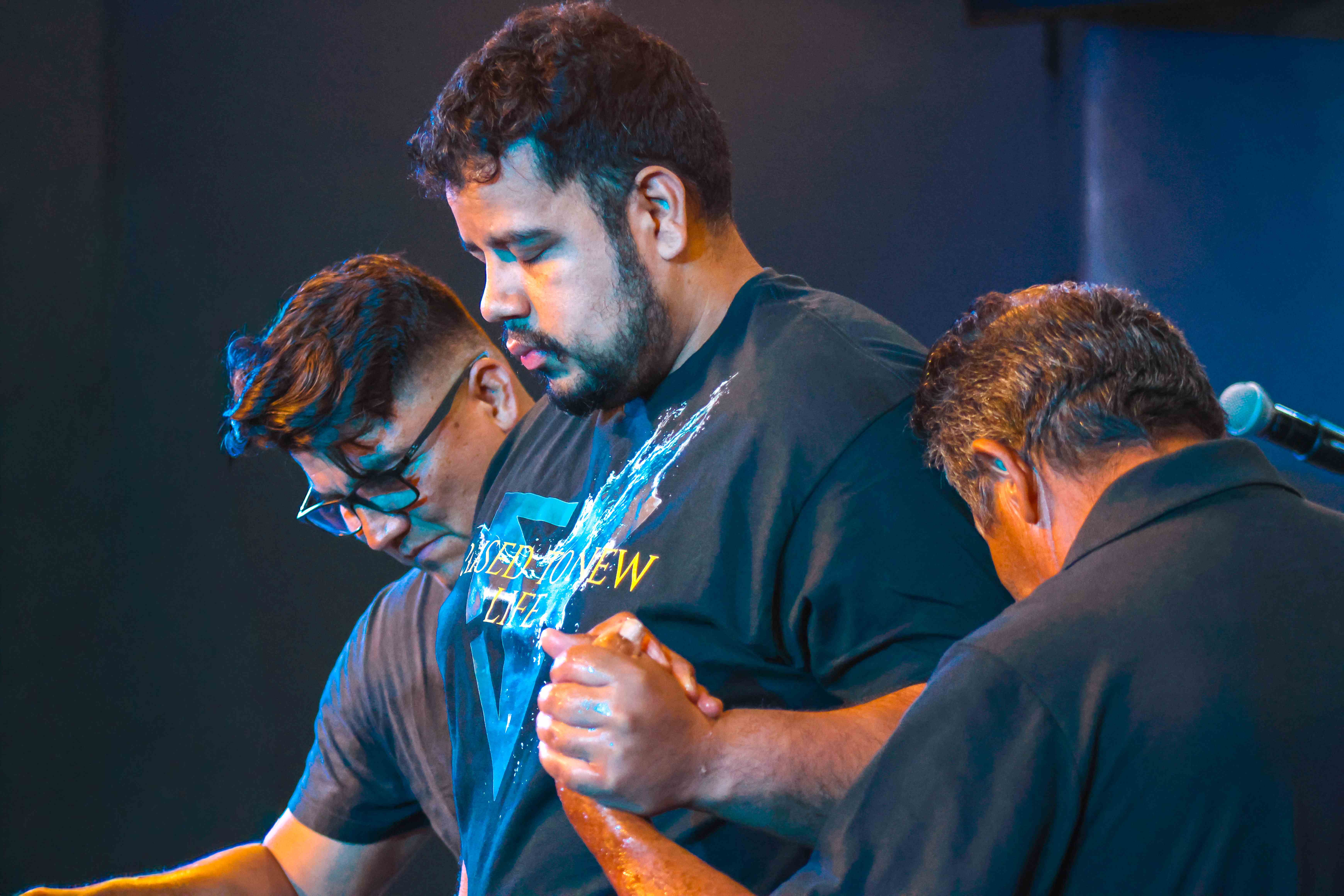 Leaders praying over a freshly baptized man in a wet Raised to New Life shirt at Ethos Church